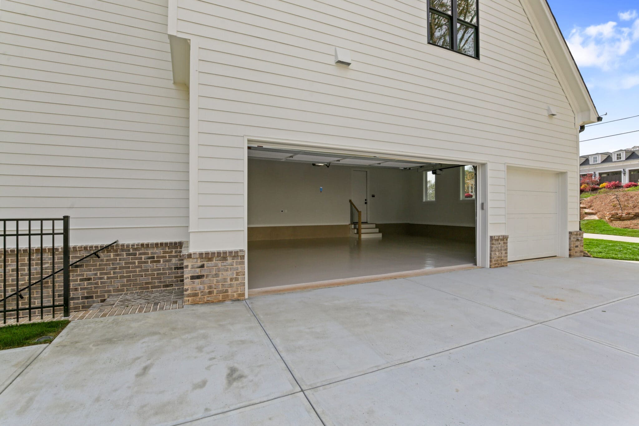 An empty garage with an open white door, attached to a beige house with brick trim. The clean concrete driveway leads up to the entrance. A small staircase is visible inside the garage.