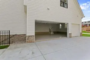 An empty garage with an open white door, attached to a beige house with brick trim. The clean concrete driveway leads up to the entrance. A small staircase is visible inside the garage.