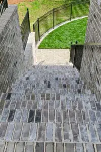 A staircase made of grey bricks descends between brick walls, leading to a sidewalk and a green grass lawn surrounded by a black metal fence.