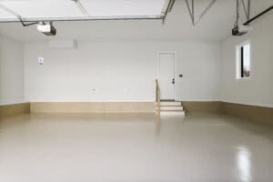 An empty, clean garage with a polished floor, white walls, and a single door leading inside. The garage door is open, and there are two windows on the right-hand side.