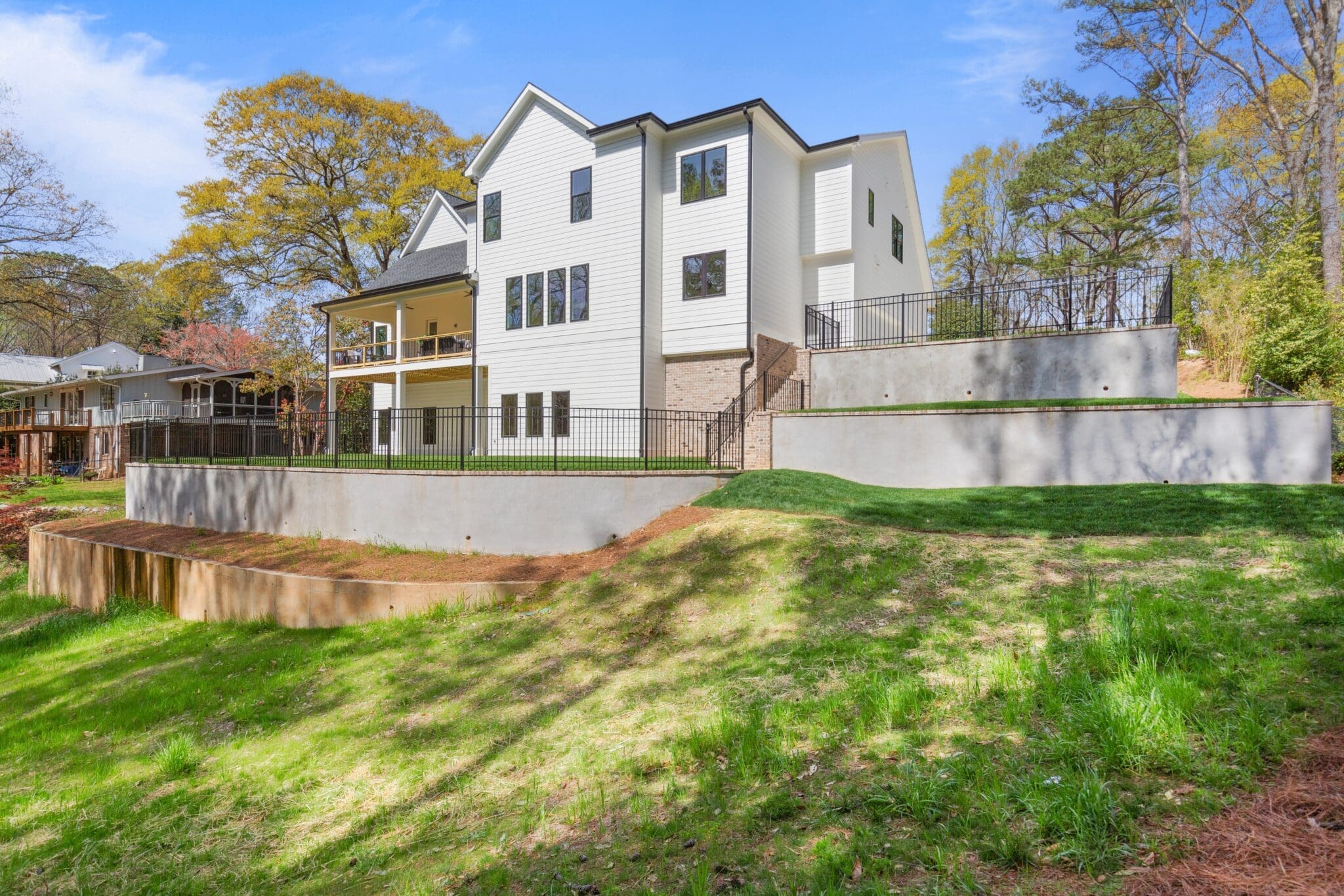 A two-story white house with black trim features a large balcony, a fenced-in yard, and is situated on a landscaped hillside. Other houses and trees are visible in the background.