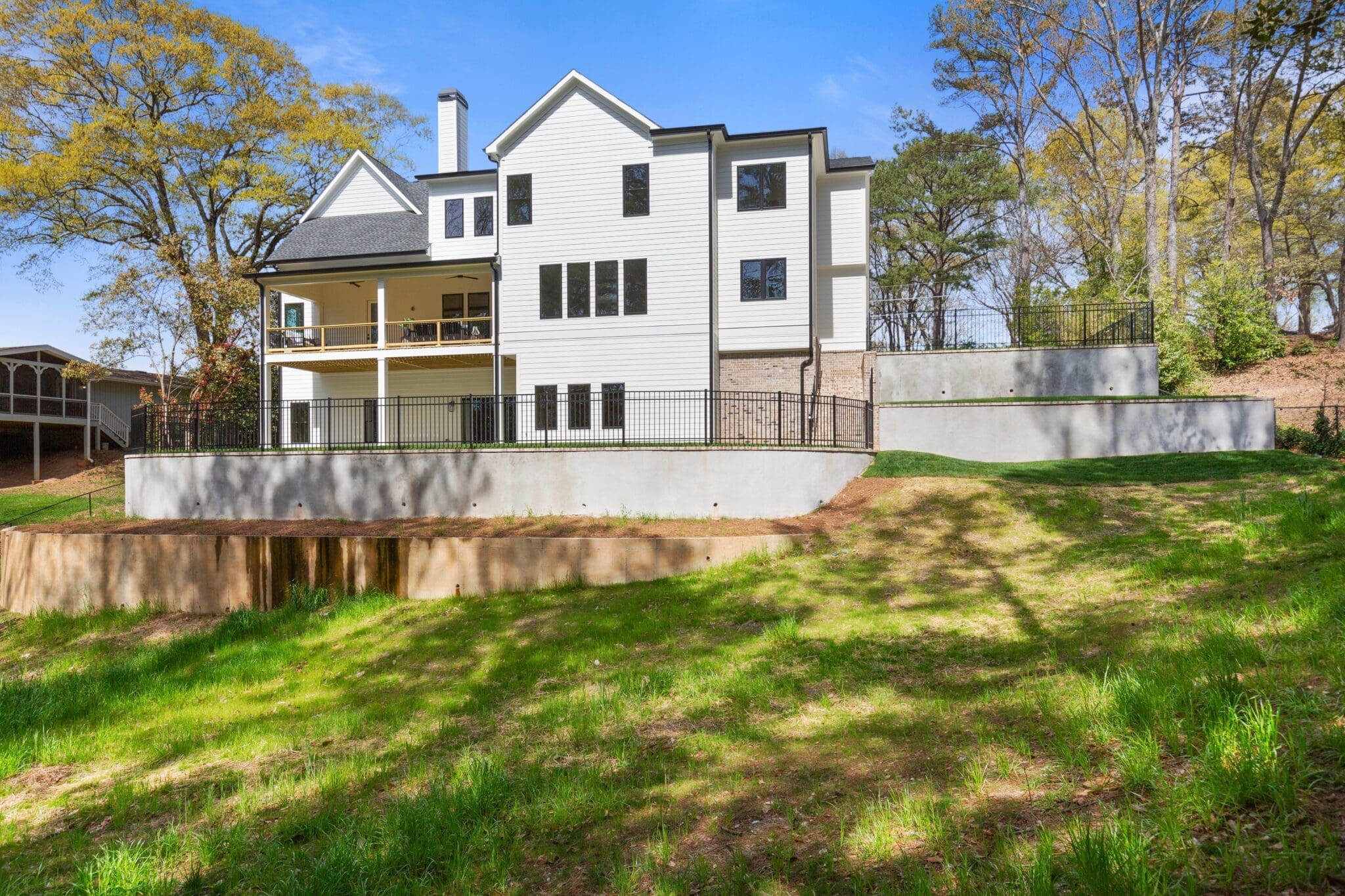 A multi-story white house with large windows and a covered porch stands on a grassy slope, surrounded by trees under a clear blue sky.