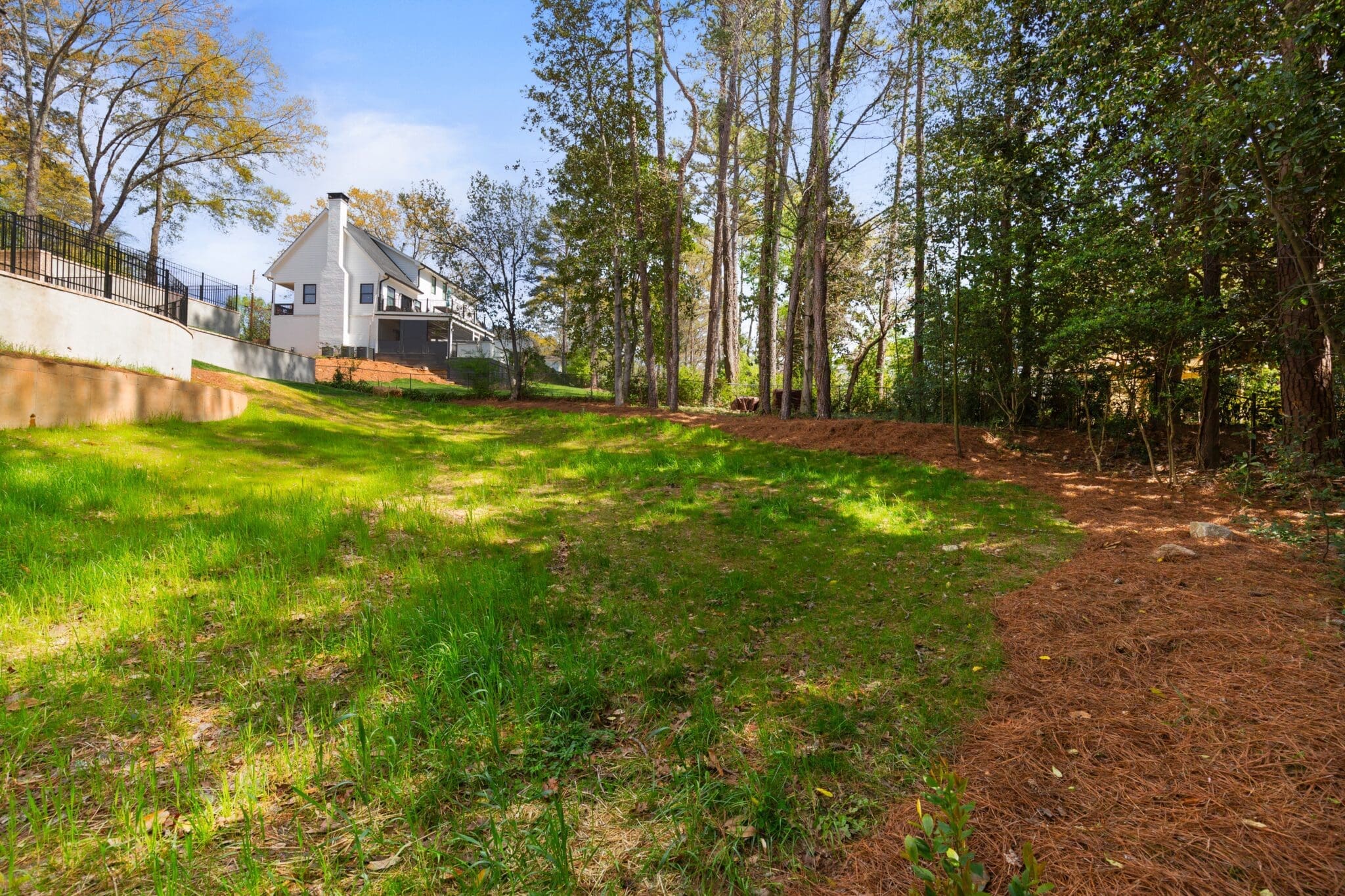 A backyard with green grass and tall trees, featuring a white house with a chimney in the background. The area is partially shaded by the trees, with a fence visible on the left side.