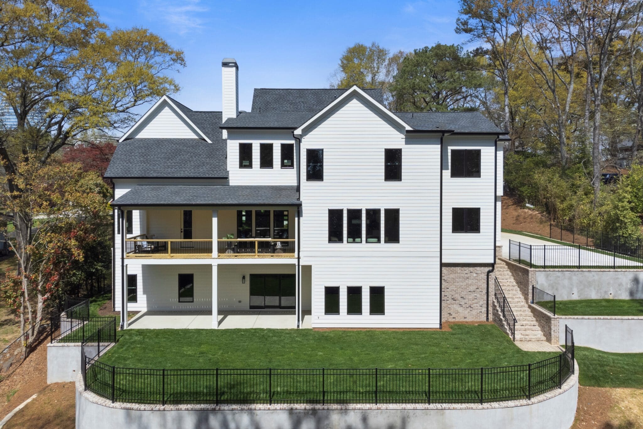 A large, modern two-story house with white exterior siding, black roof, multiple windows, and a covered balcony. The backyard is fenced and features a well-maintained lawn and concrete pathways.