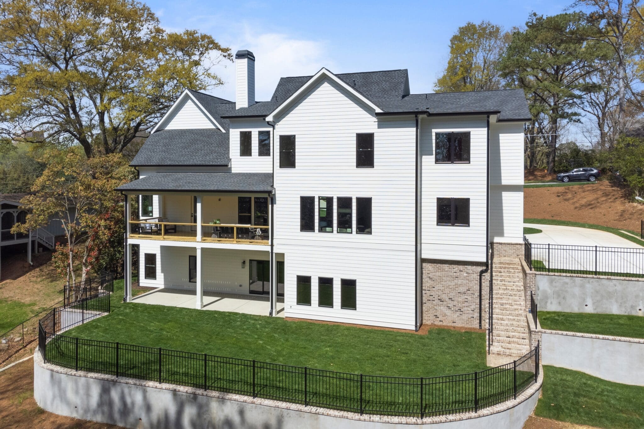 A large, two-story modern house with white siding, black shutters, and multiple windows. The house has a spacious green lawn, a fenced yard, and a stone retaining wall on one side.