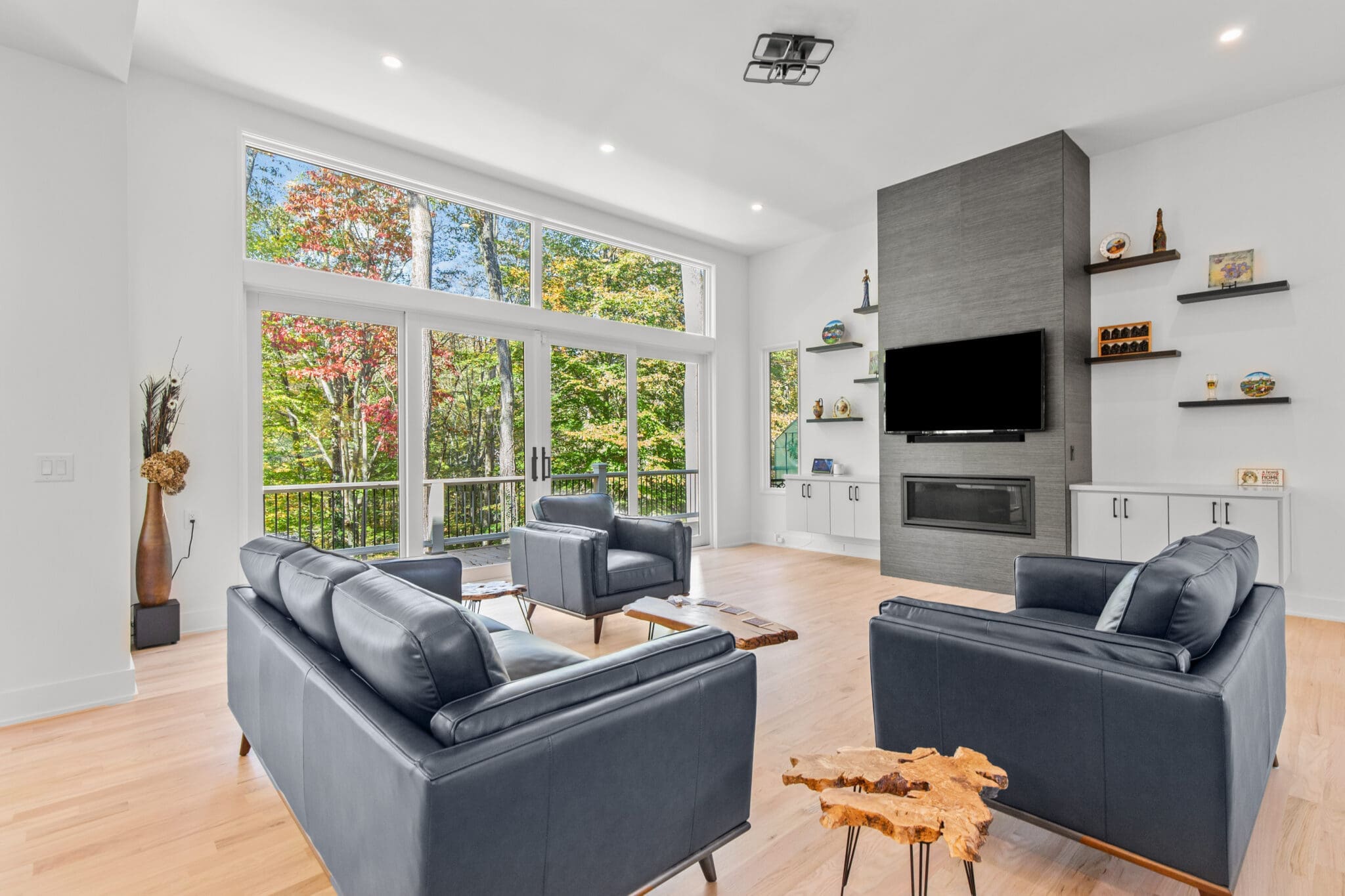 A modern living room with large windows, blue sofas, a wooden coffee table, built-in cabinets, and a wall-mounted TV above a fireplace. The windows show greenery outside.