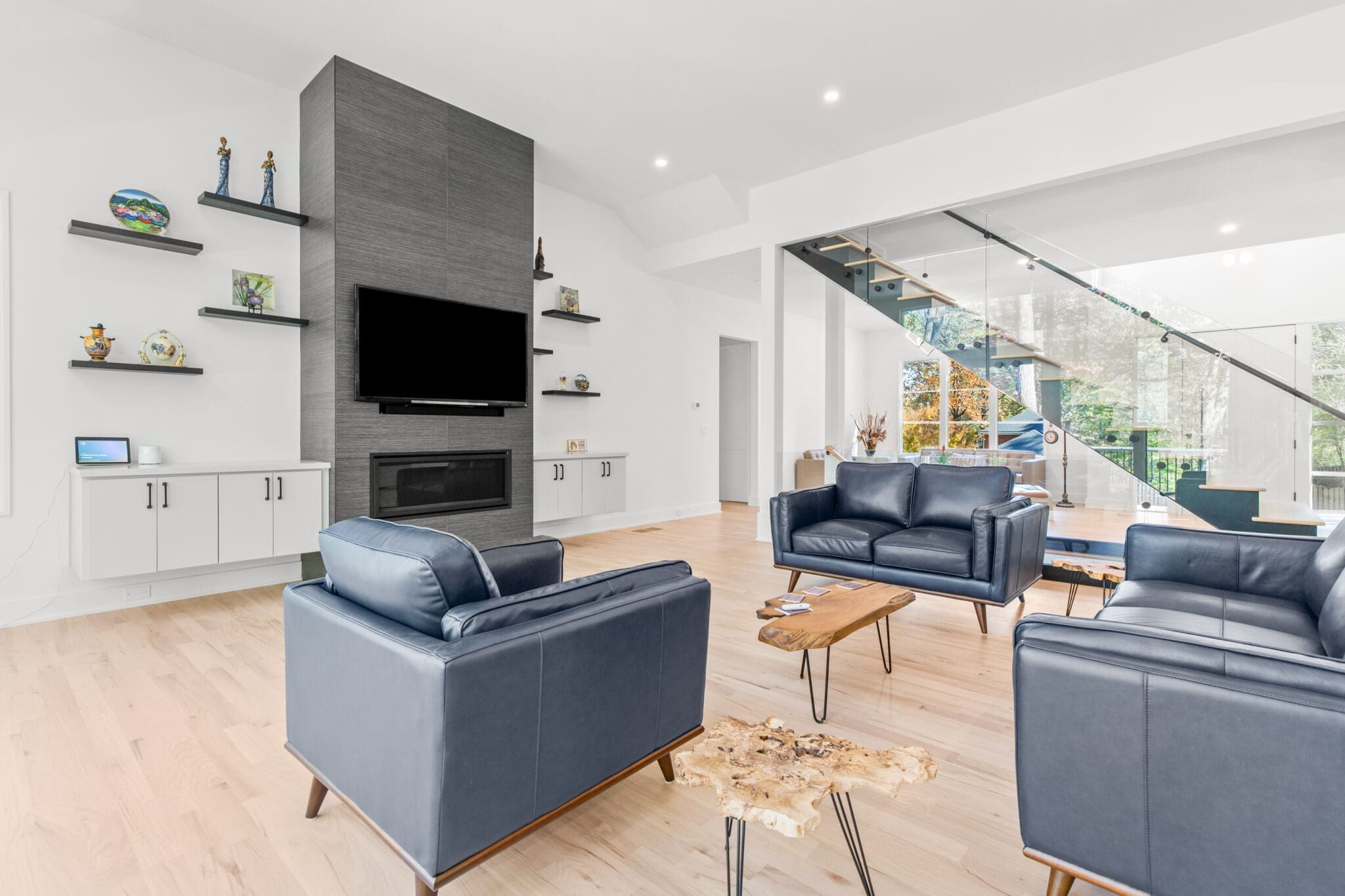 Modern living room with dark blue leather sofas, a sleek fireplace with a flat-screen TV above, floating shelves with decor, a wooden coffee table, and a glass staircase leading to the upper floor.