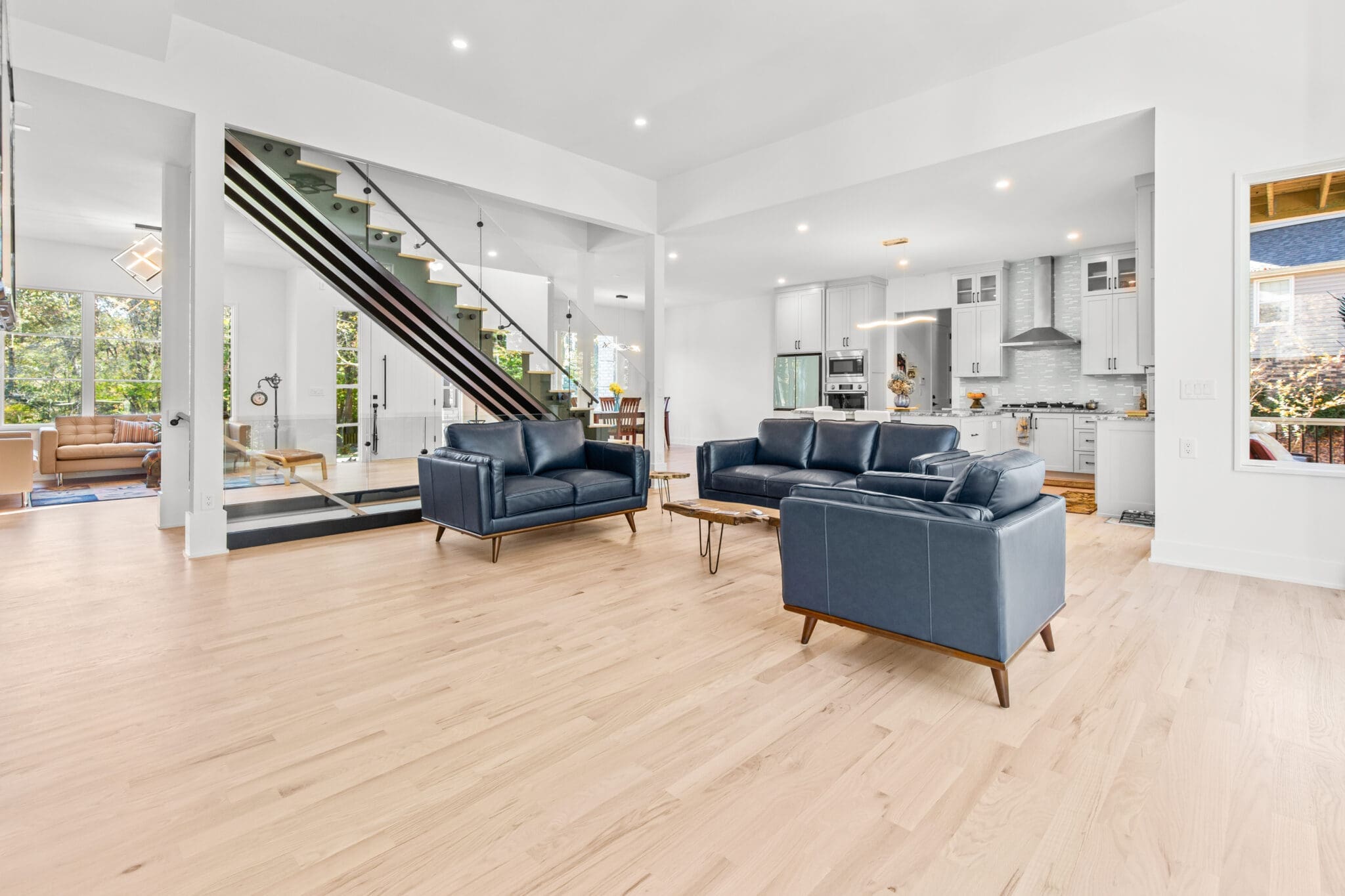 A spacious, modern living area with two blue sofas, a wooden coffee table, a staircase with glass railing, and an adjacent kitchen with white cabinetry and stainless steel appliances.