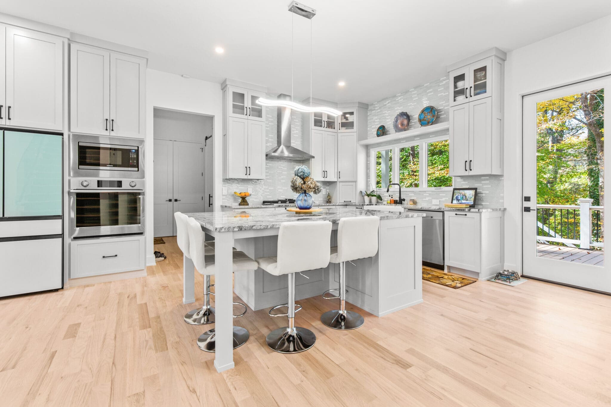 A modern kitchen features white cabinetry, stainless steel appliances, a marble island with three white barstools, wooden floors, and large windows with a view of greenery.