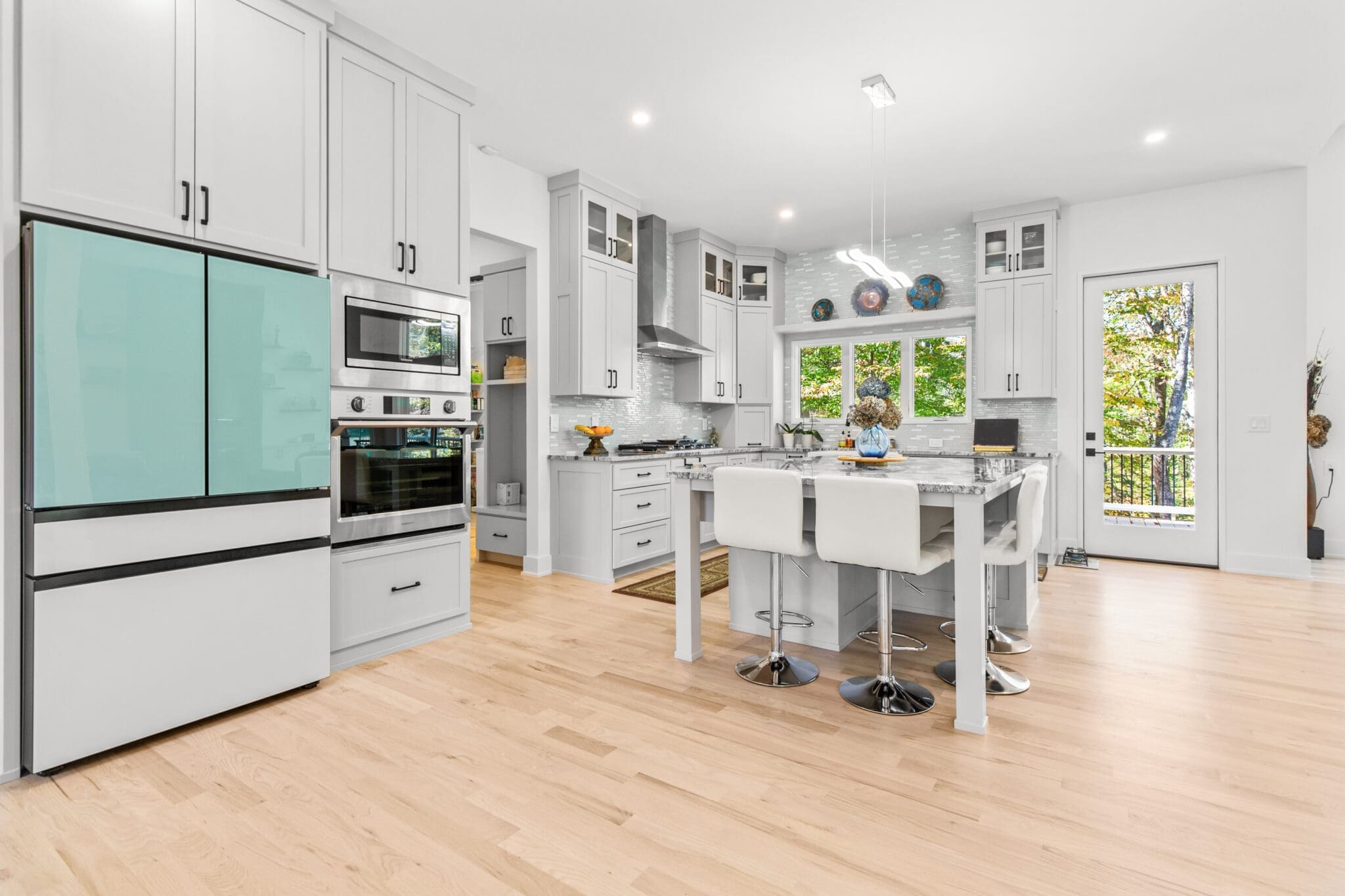 Modern kitchen with white cabinetry, stainless steel appliances, a center island with stools, and wood flooring. Large windows and a glass door provide ample natural light.
