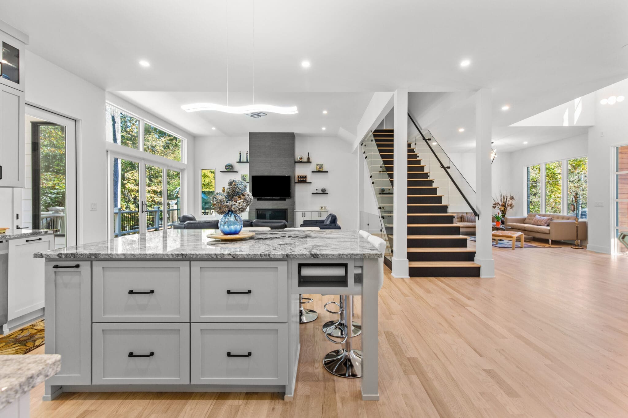 Modern open-plan kitchen and living area with light wood flooring, large kitchen island, white cabinets, and a black staircase leading to the second floor, illuminated by natural light from large windows.