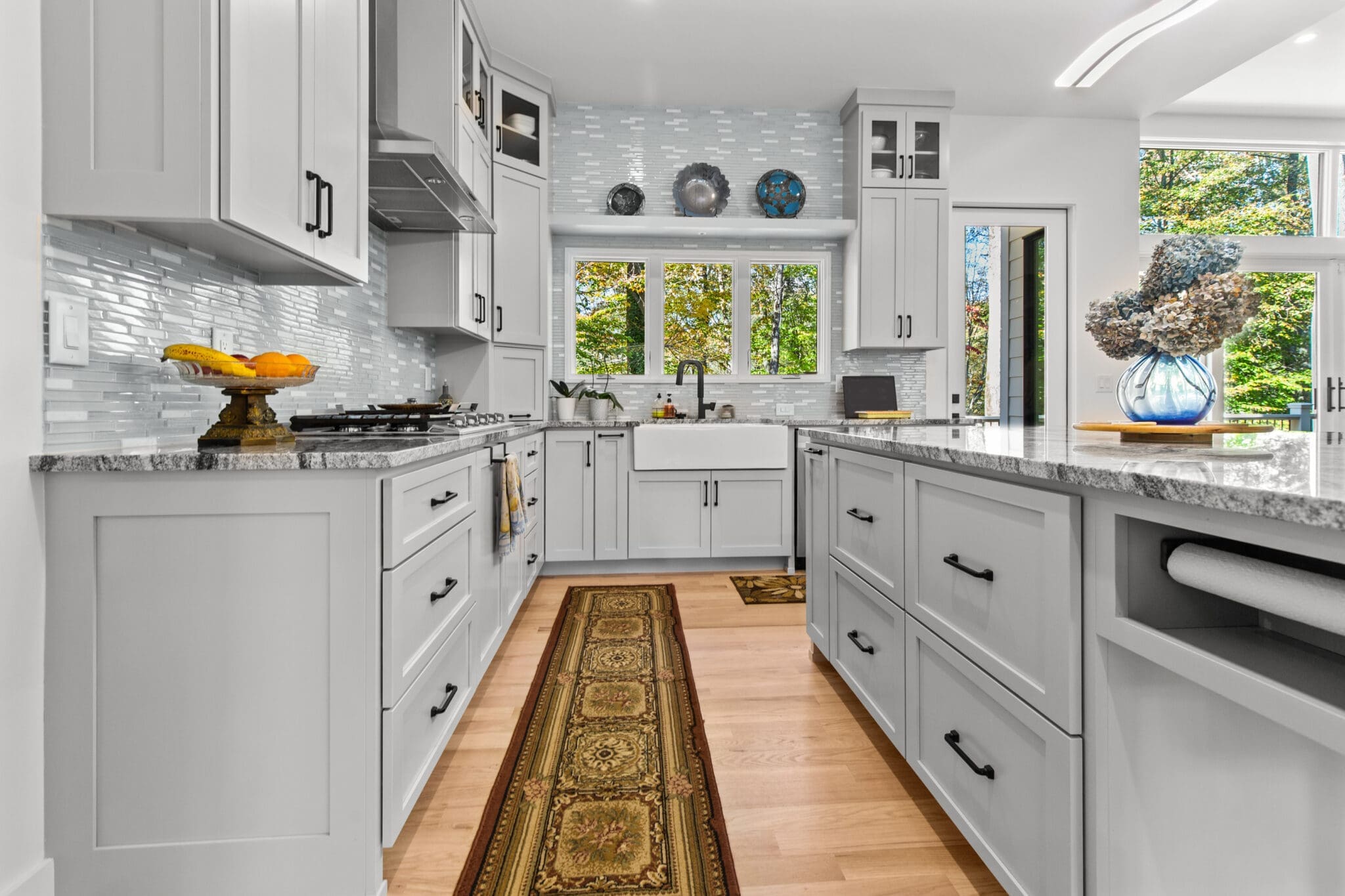 A modern kitchen with white cabinetry, granite countertops, stainless steel appliances, and a central island. A patterned rug runs along the wooden floor, and a vase with flowers is on the island.