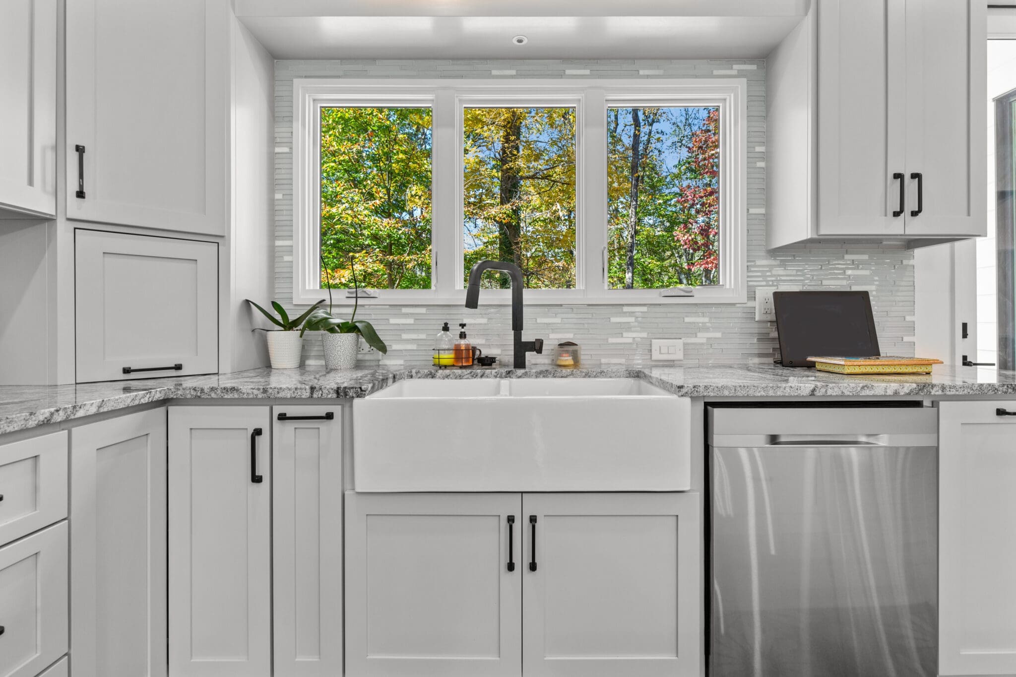 A modern kitchen with white cabinets, a farmhouse sink, a black faucet, and a stainless steel dishwasher. There's a window with a view of trees, and potted plants and kitchen items on the counter.
