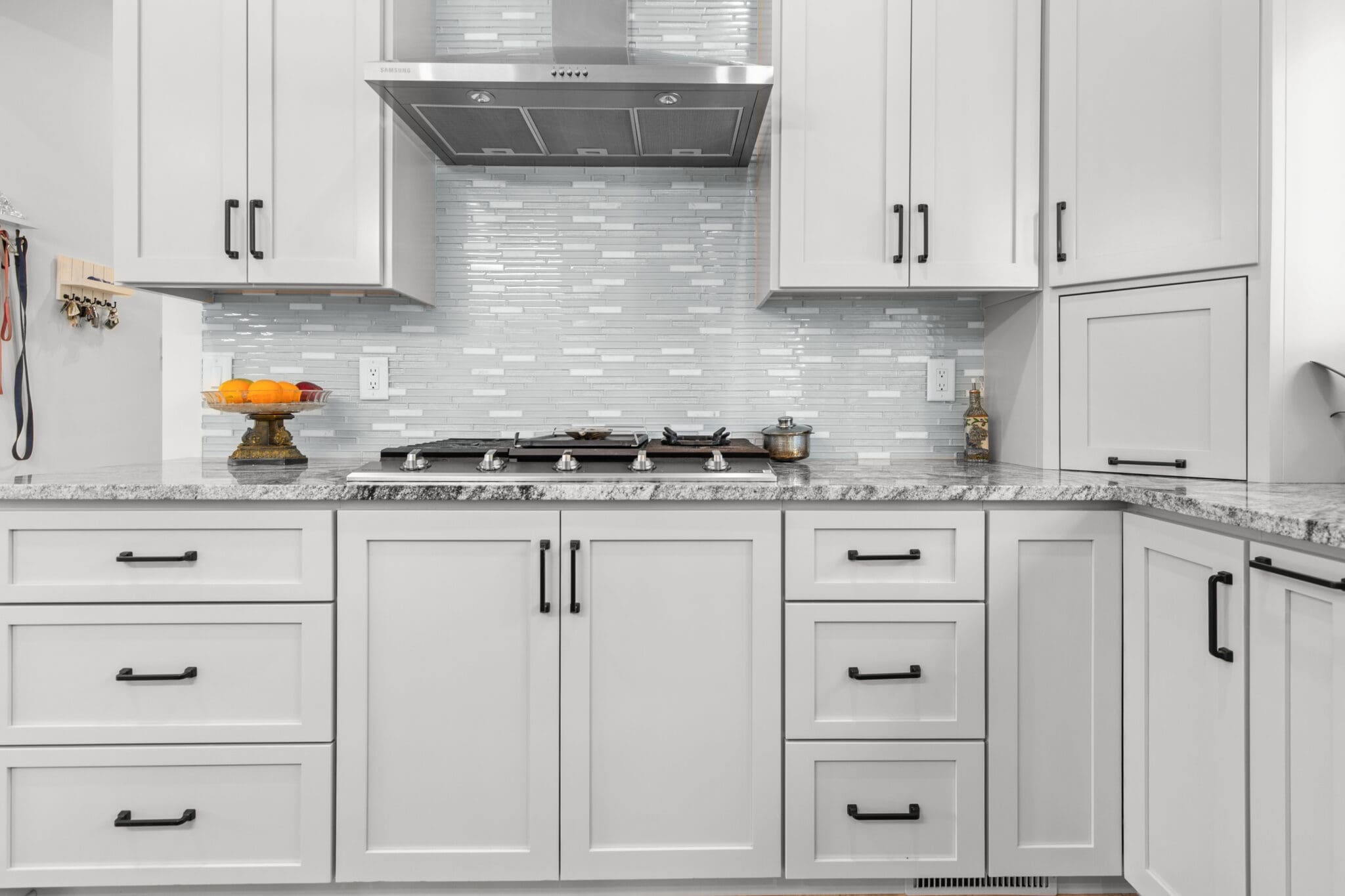 A modern kitchen with white cabinets, a stainless steel range hood, and black hardware. The backsplash features horizontal white and gray tiles, and there is a granite countertop.