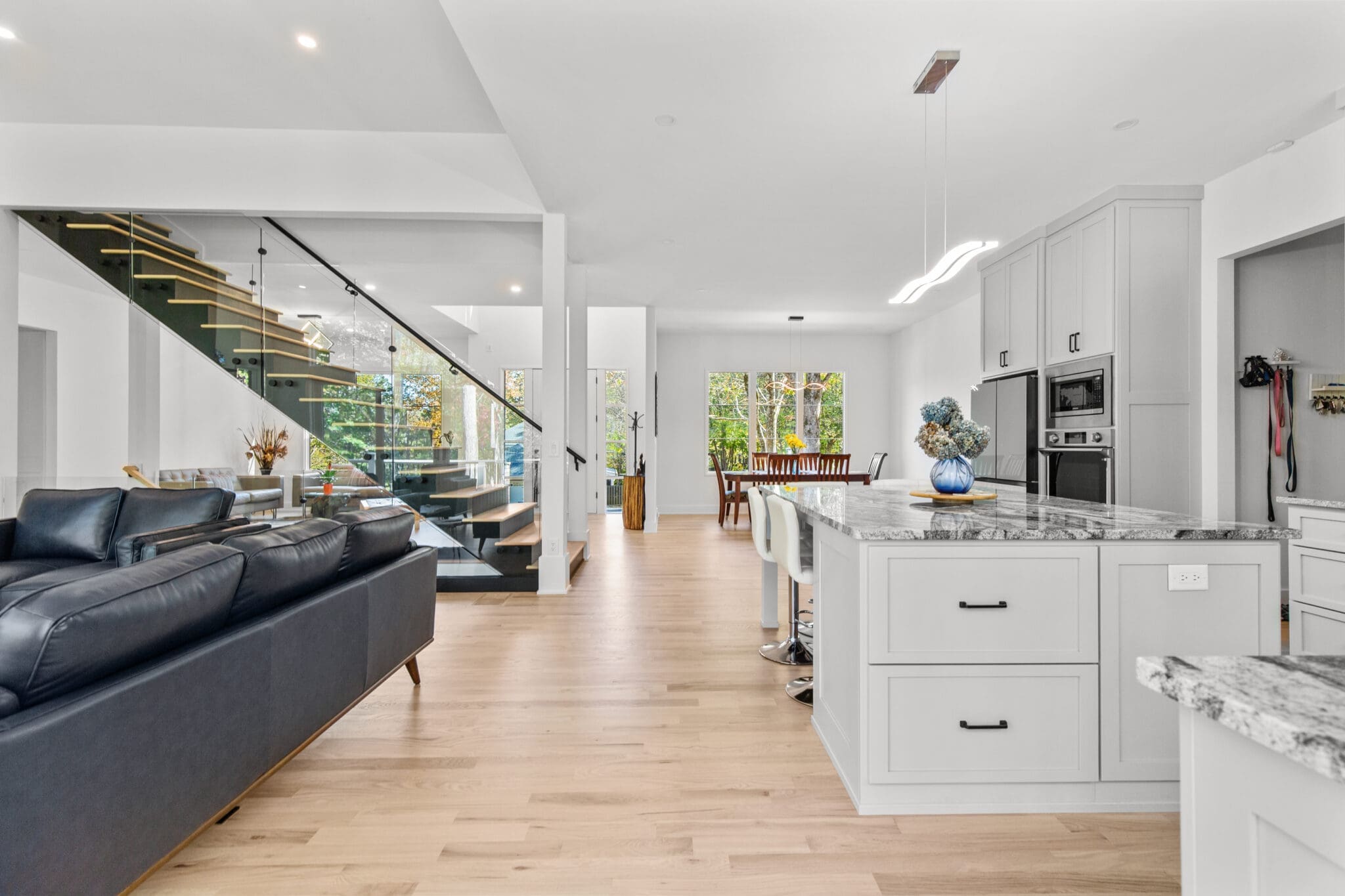 A modern open-concept living space with a kitchen island, dining area, black staircase with glass railing, and a black couch. Natural light streams through large windows, and the space has white walls and wooden floors.