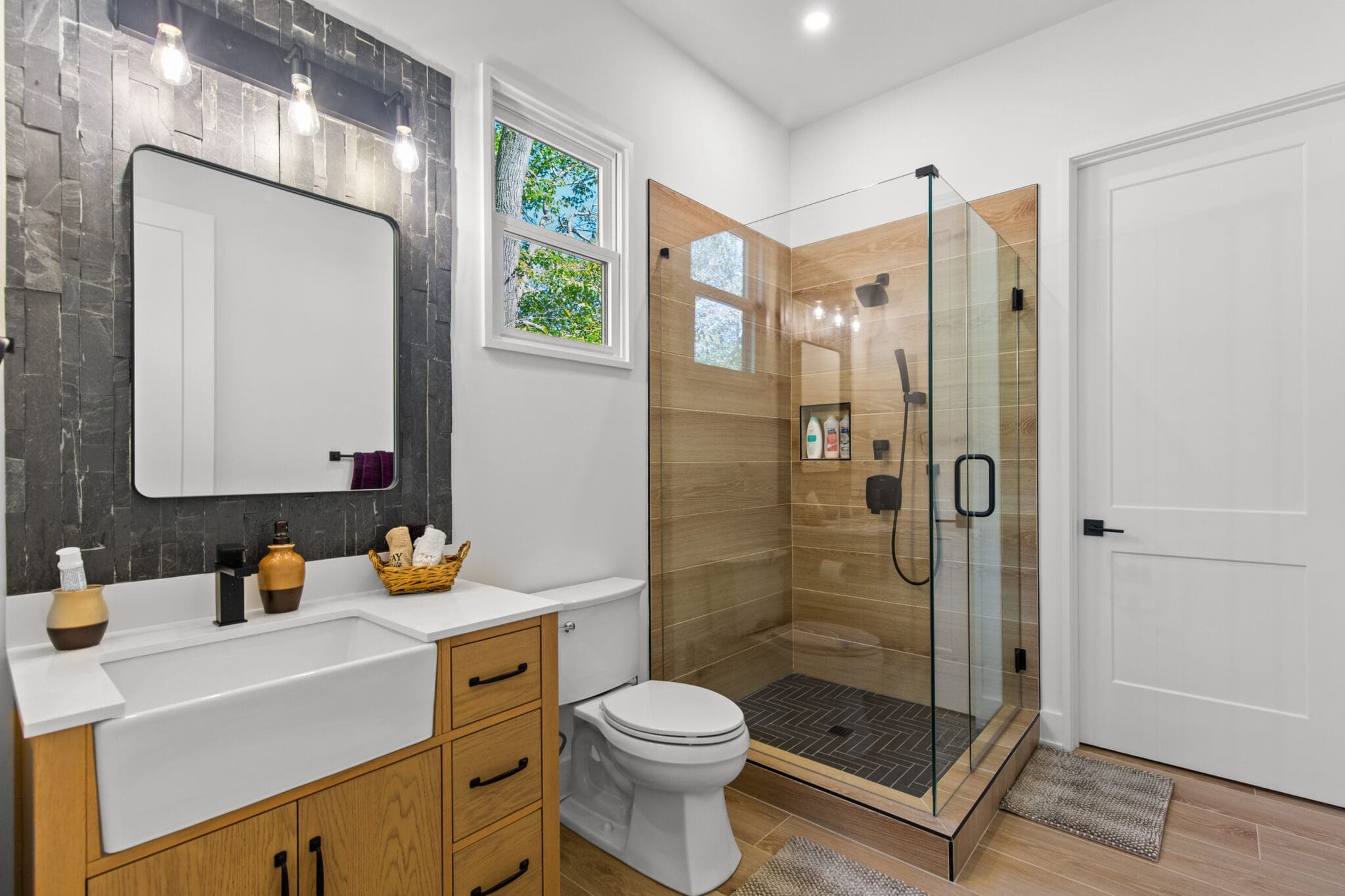 Modern bathroom with a wooden vanity, white sink, and toilet. A glass-enclosed shower is next to a white door. A small window and sconce lights brighten the space.