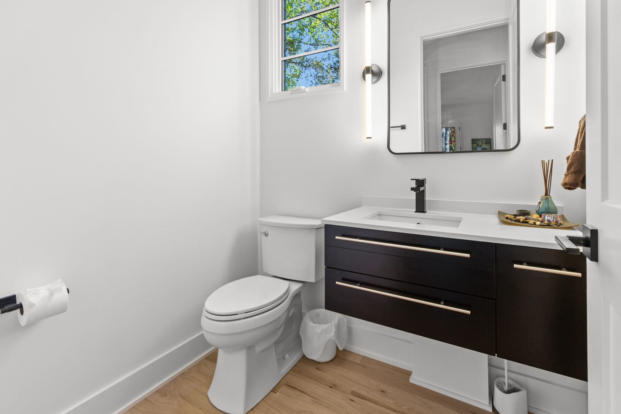A modern bathroom with a white toilet, dark wood vanity with sink, large mirror, two vertical light fixtures, and a window above. There are wooden floors, a trash bin below the sink, and a toilet paper holder on the left wall.