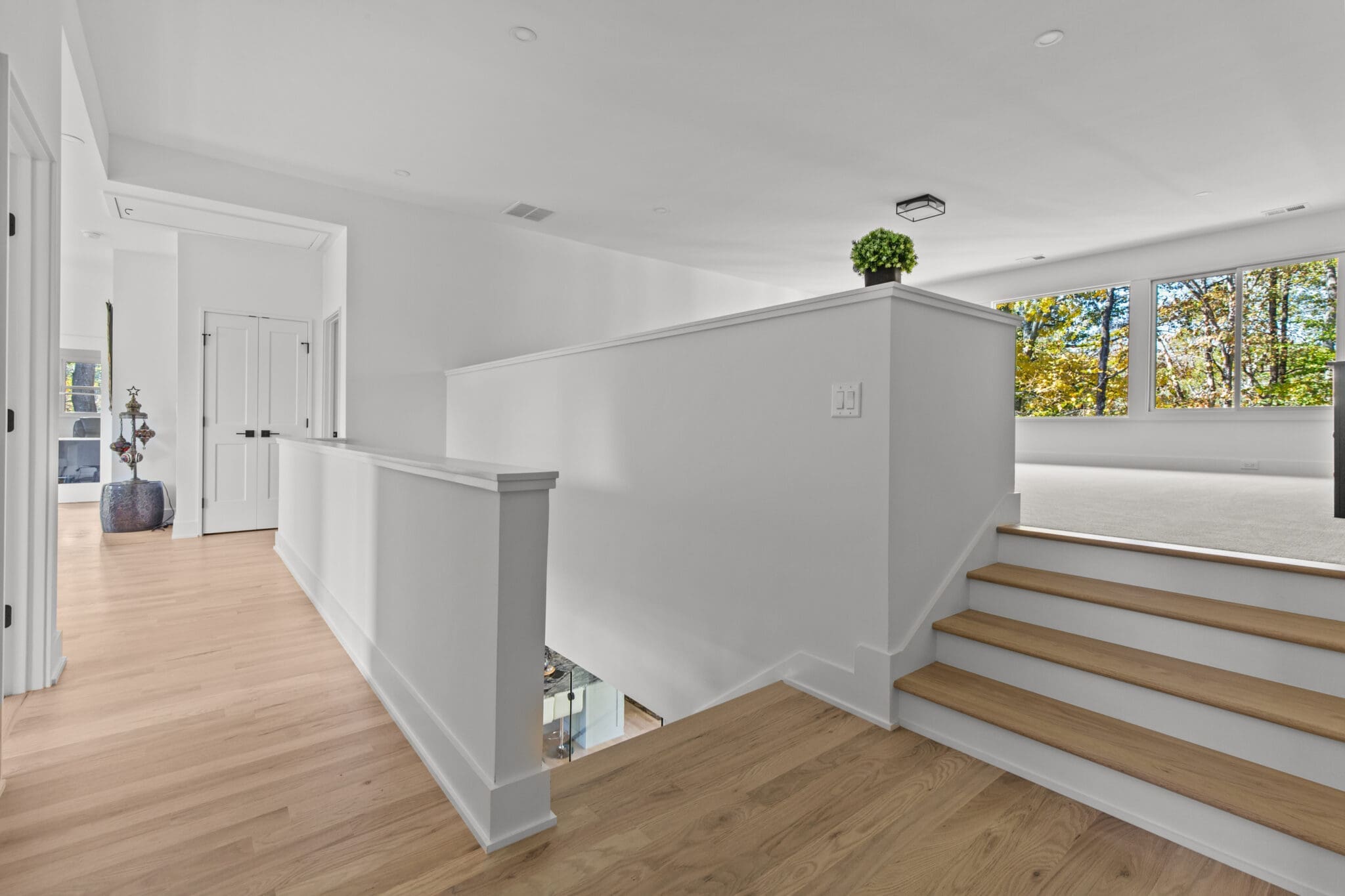 A modern, white-painted hallway with wooden flooring and a partial wall overlooking a spacious, bright room. Wooden stairs lead up from the lower level. Green plants and large windows are visible.