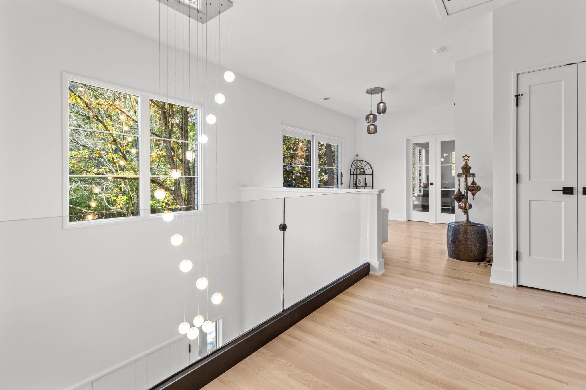 Bright hallway with wooden flooring, modern hanging light fixtures, and large windows. The space features minimalistic decor and a glass barrier along the stairwell.
