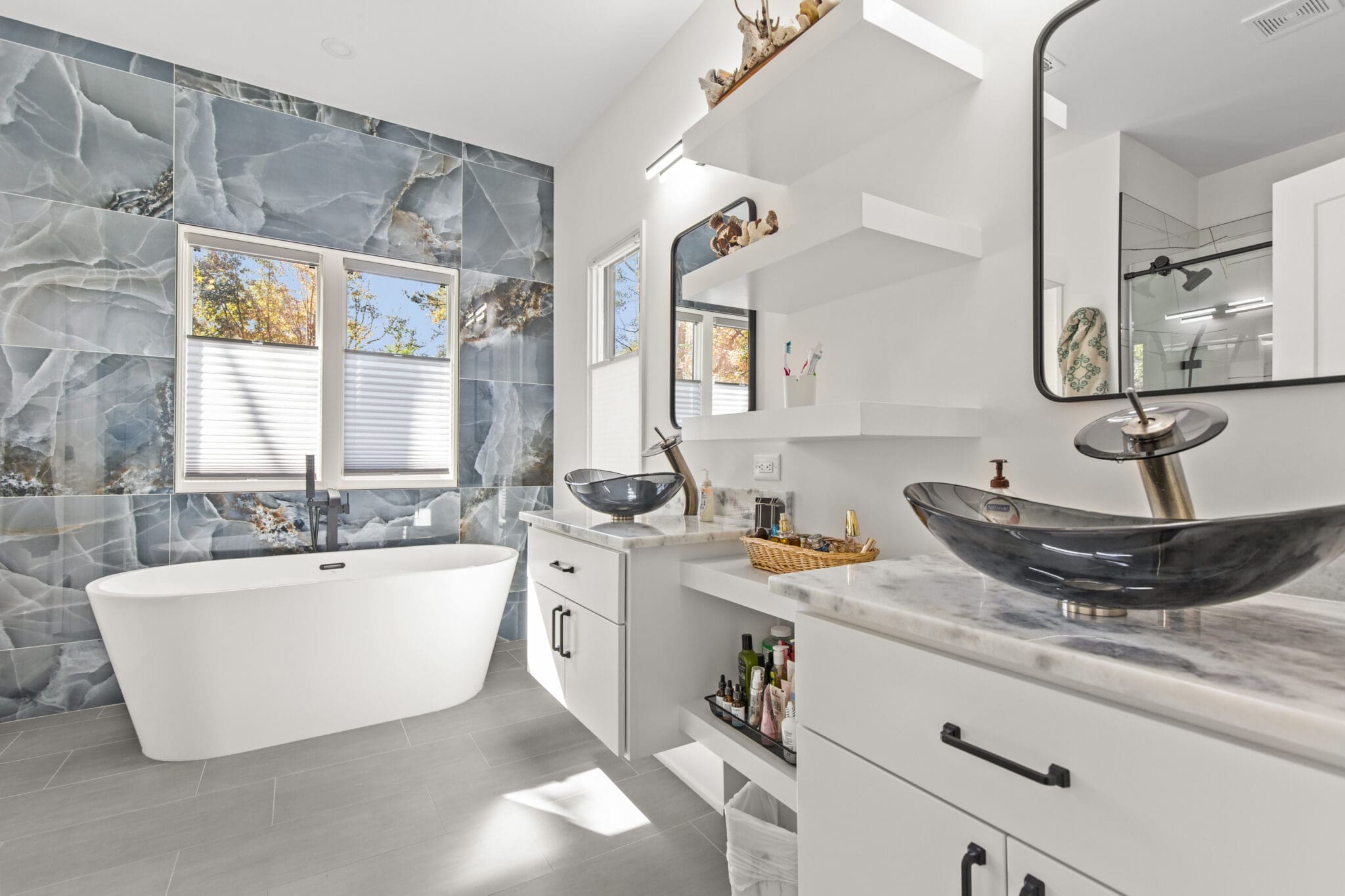 A modern bathroom with a freestanding bathtub, two black vessel sinks on a marble countertop, white cabinets, and a large blue-patterned accent wall. A window with frosted blinds lets in natural light.
