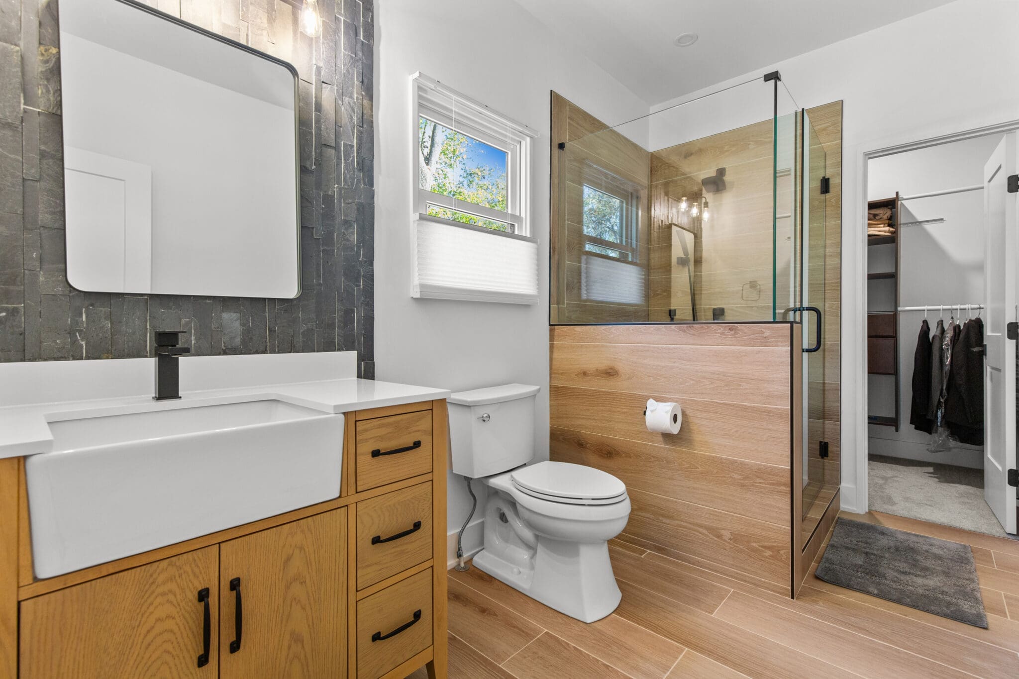 A modern bathroom with a wooden vanity, white sink, toilet, and a glass-enclosed shower. There's a small window near the shower, and an open door leads to a walk-in closet with clothes hanging.