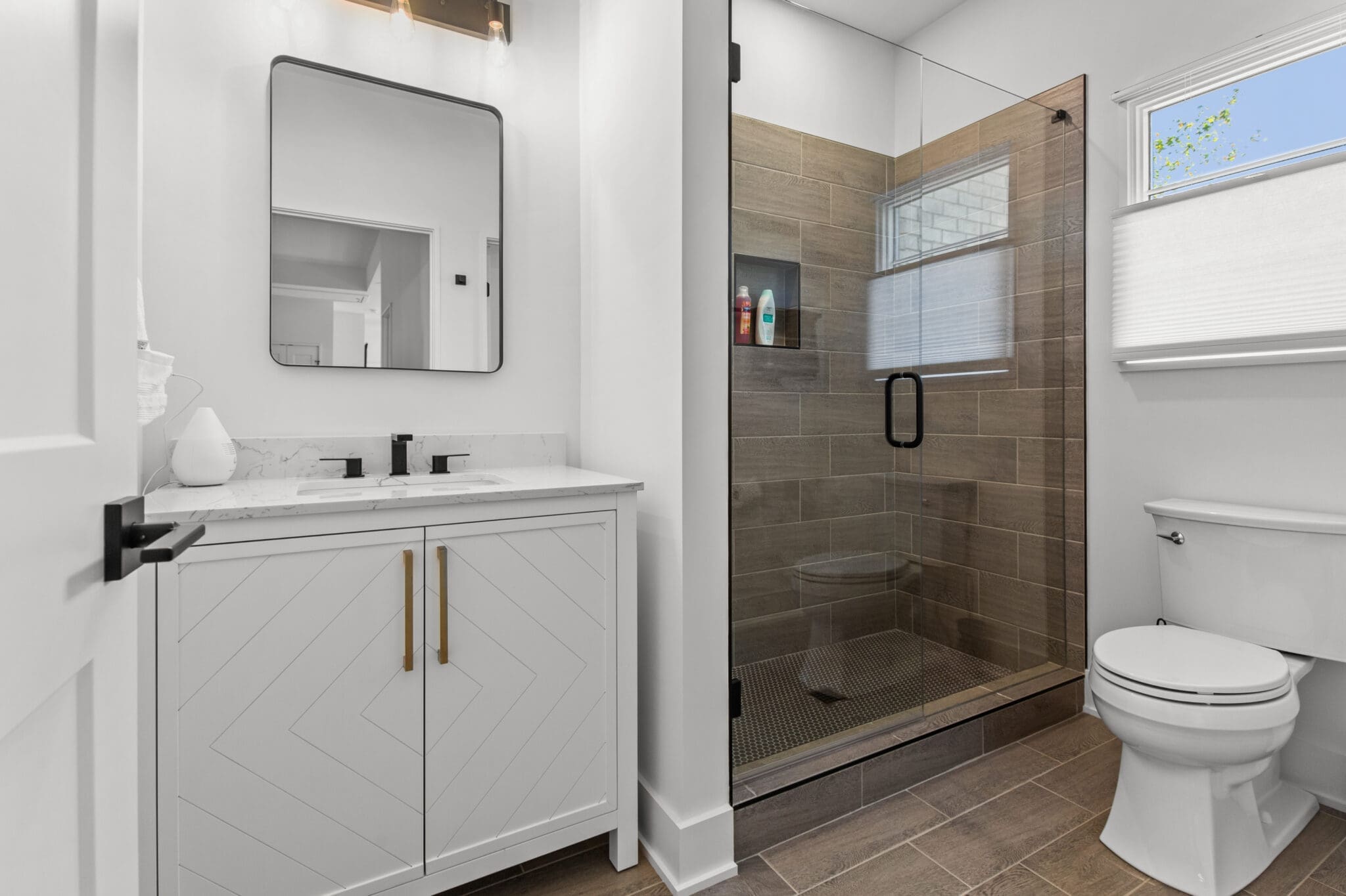 Modern bathroom with a white vanity and dual faucets, a rectangular mirror, a glass-enclosed shower, and a white toilet. The shower has built-in shelves and brown tiles.