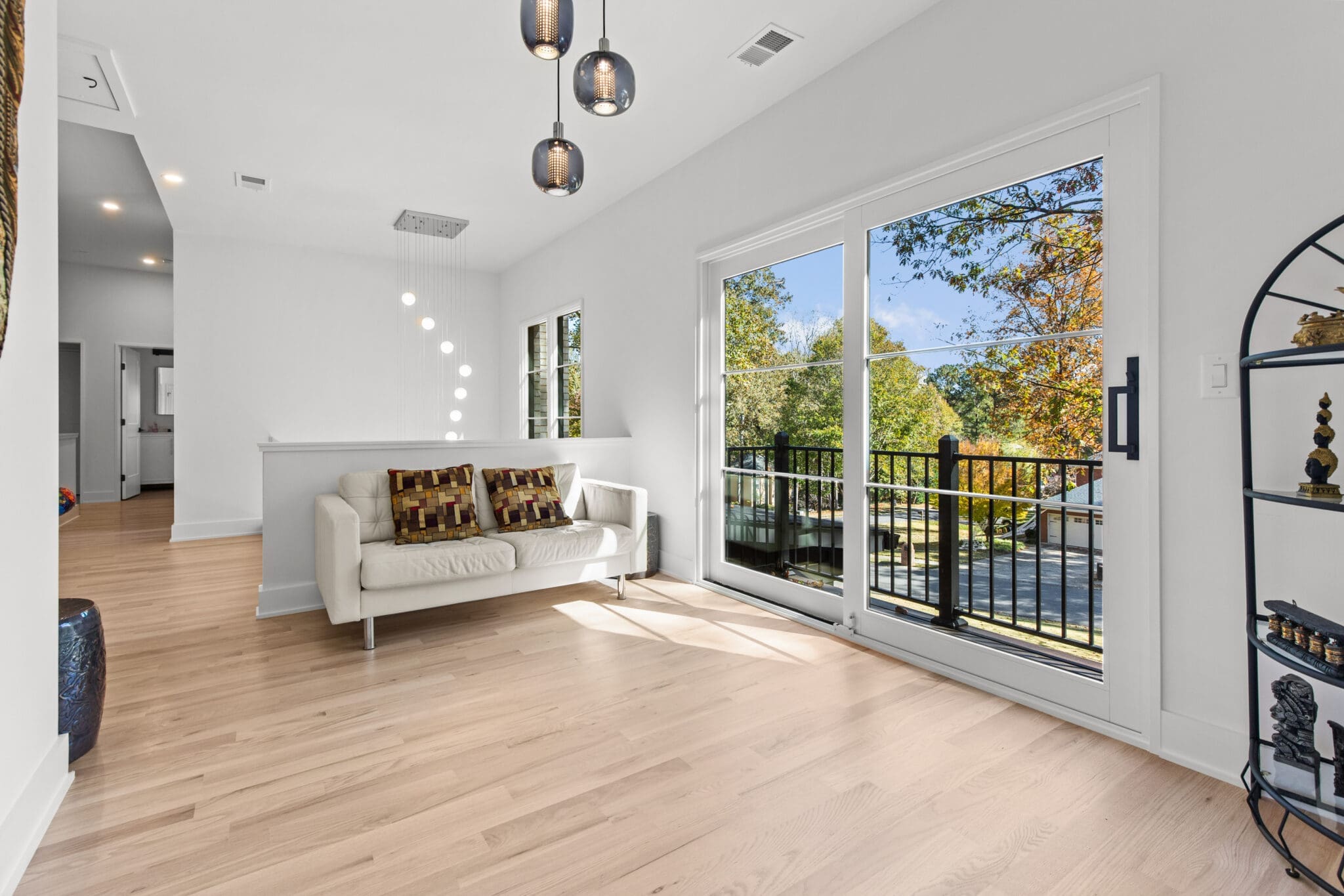A bright room with a white sofa, decorative pillows, hanging lights, a sliding glass door leading to a balcony, and a black shelf. The room has light wooden floors and white walls.