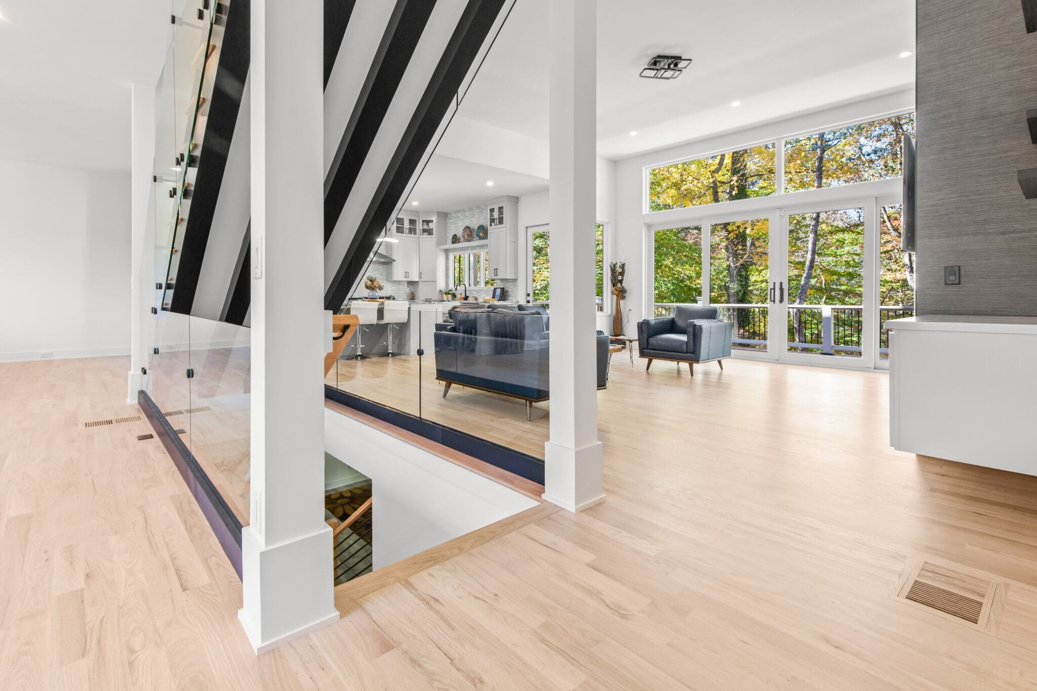 Modern open-concept living area with hardwood floors, large windows, and a staircase with black and white railing. The space includes blue sofas, a dining area, and a view of outdoor greenery.