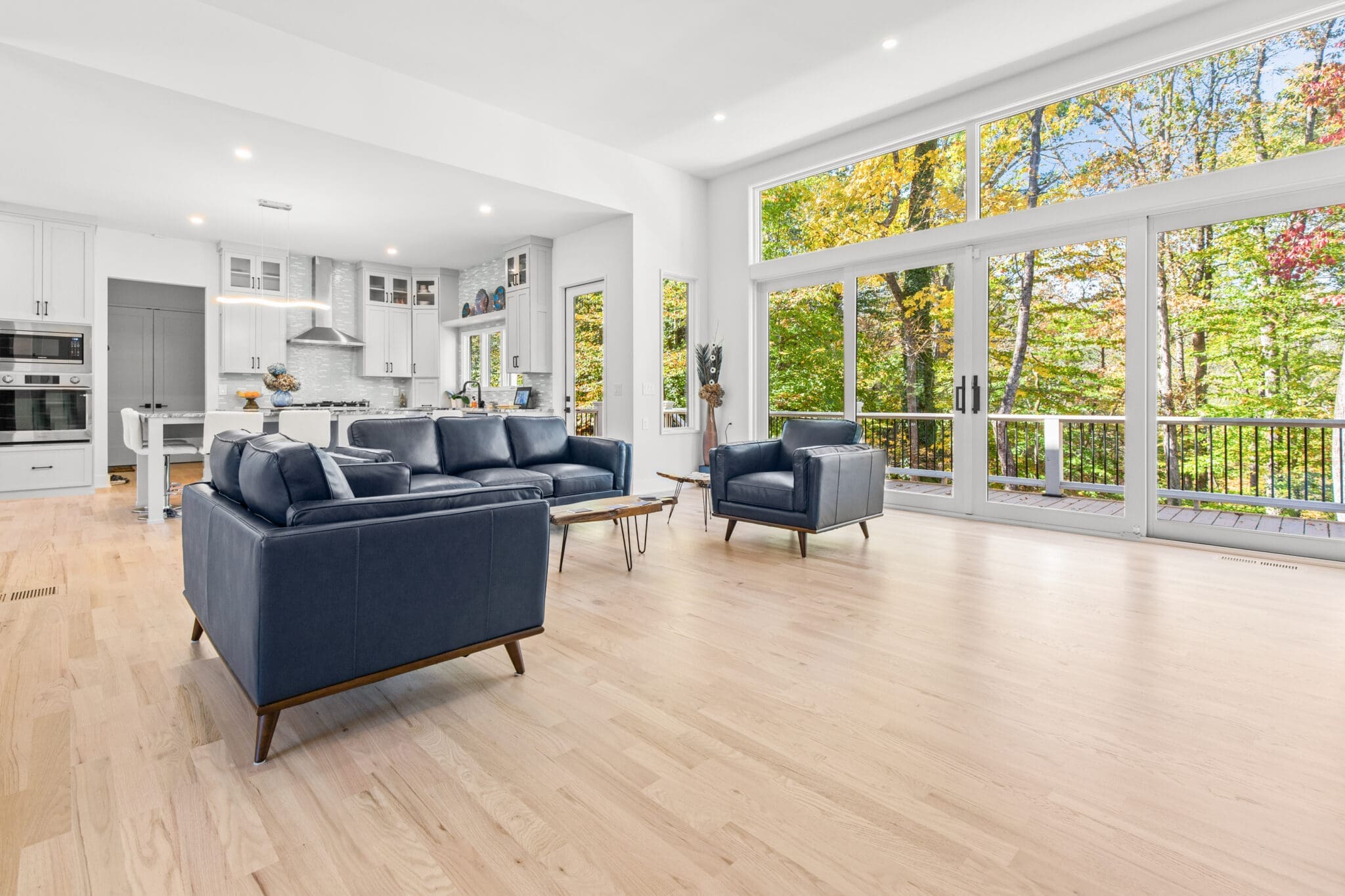 A modern living room features navy blue furniture, floor-to-ceiling windows, light hardwood flooring, and an open view of a forested area. A kitchen with white cabinets is visible in the background.