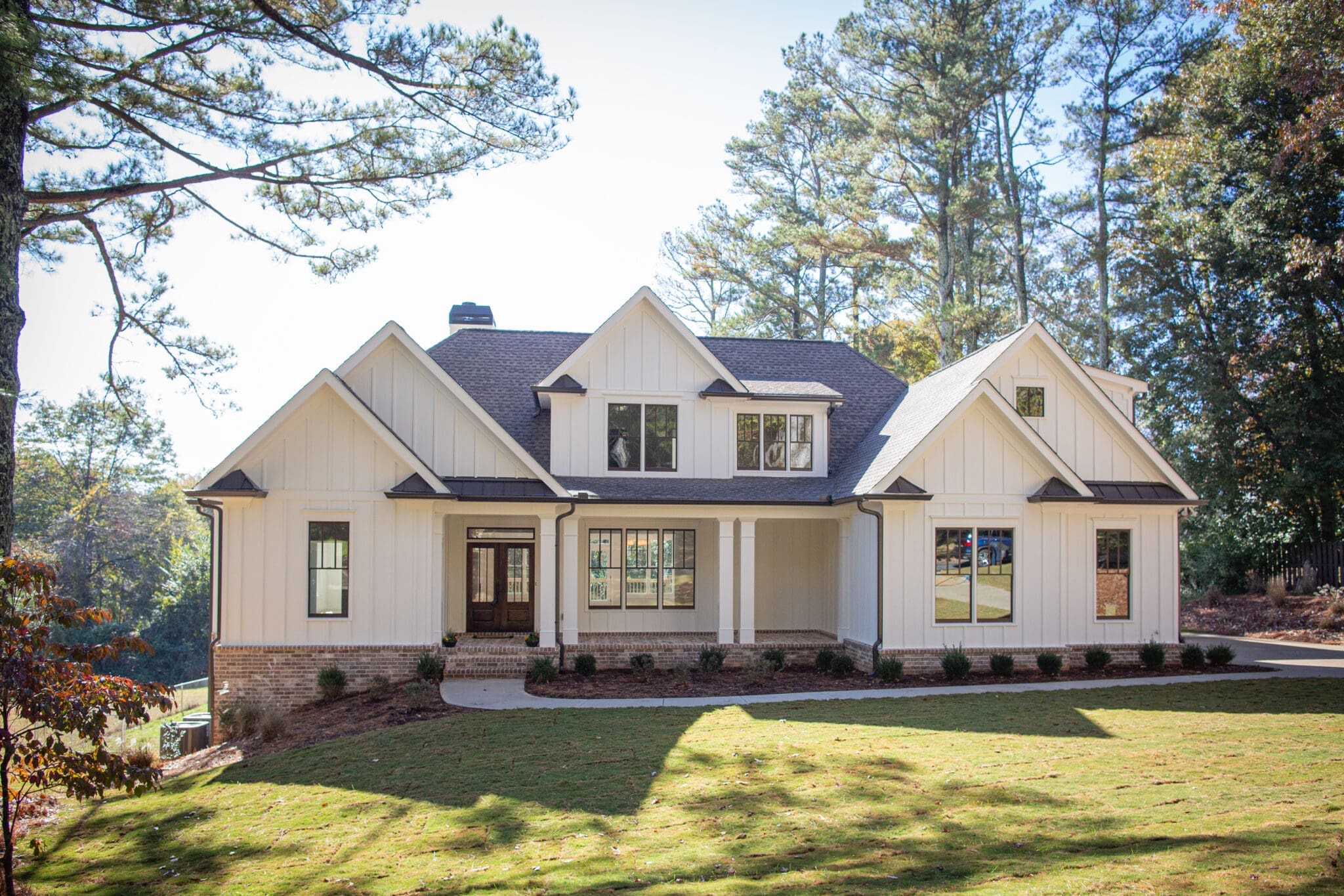A white two-story house with a dark gray roof, gabled dormers, and a covered front porch, set on a spacious lawn and surrounded by tall trees.
