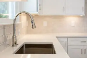 Close-up of a modern kitchen sink with a stainless steel faucet set in a white countertop, framed by white cabinetry and neutral-toned backsplash tiles.