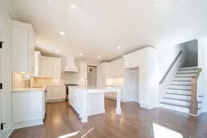 Bright, modern kitchen with white cabinetry, a central island, stainless steel appliances, hardwood floors, and adjacent staircase leading to the upper floor.