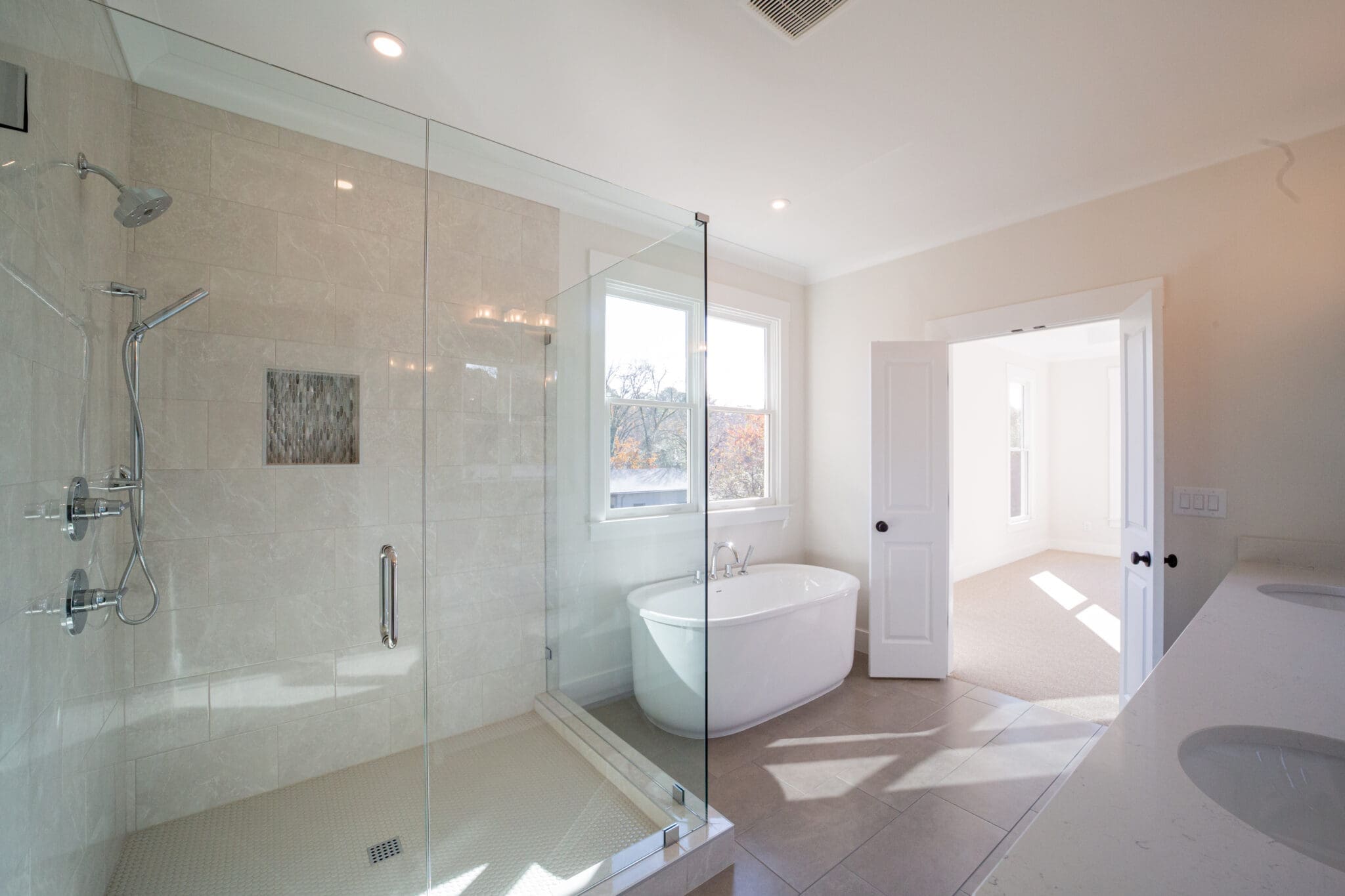 A modern bathroom features a glass-enclosed shower, a freestanding bathtub, dual sinks, and large windows letting in natural light.