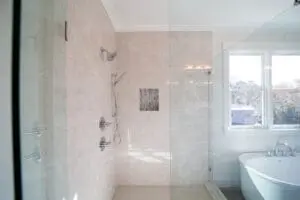 Brightly lit bathroom featuring a glass-enclosed shower with multiple showerheads, a tiled wall, and a standalone bathtub next to large windows.