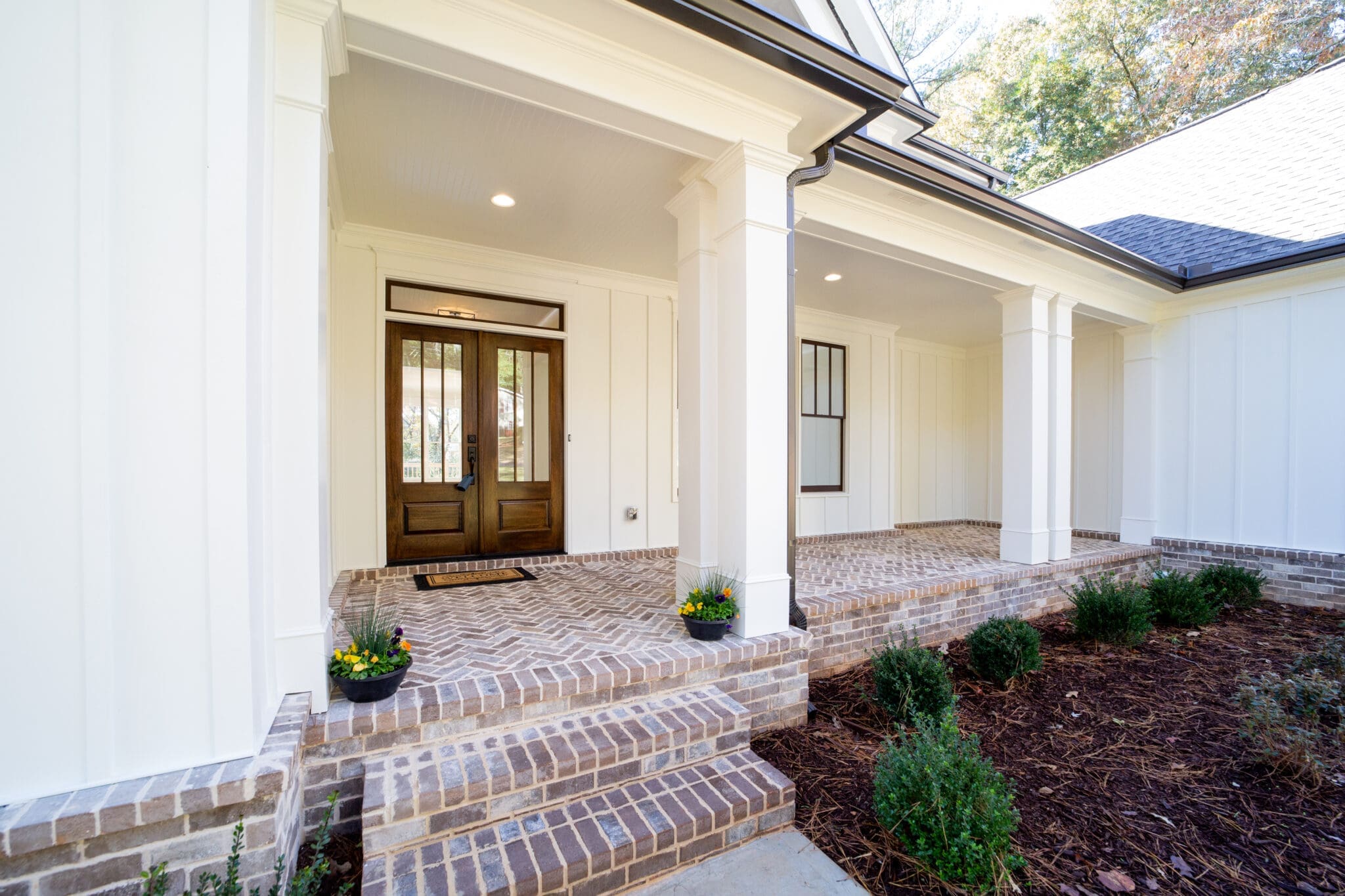 Front entrance of a house with a brick porch, double front doors, and white columns. Flower pots and small bushes are placed around the entrance.