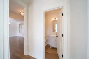 A hallway with wooden flooring leads to a small bathroom with a sink and mirror. Adjacent to the bathroom, there is a spacious, empty room visible.