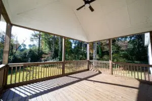 A spacious outdoor deck with a wooden floor and railing, surrounded by trees. The deck has a white, vaulted ceiling with an overhead fan.