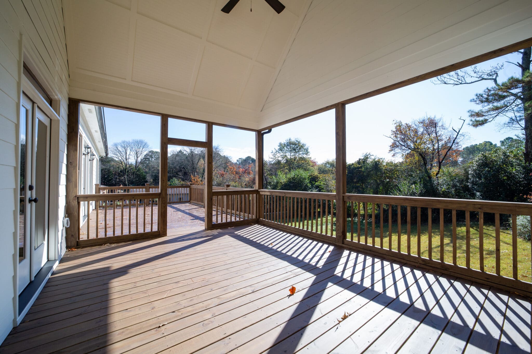 A covered, screened porch with wooden flooring overlooks a grassy backyard with trees in the distance. Sunlight casts shadows through the railings on the floor.