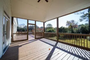 A covered, screened porch with wooden flooring overlooks a grassy backyard with trees in the distance. Sunlight casts shadows through the railings on the floor.