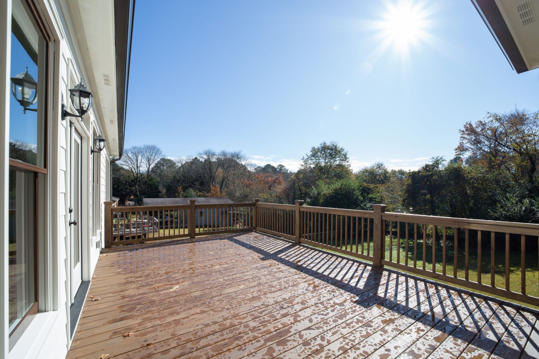A sunlit wooden deck attached to a house, with a railing overlooking a grassy yard and trees under a clear blue sky.