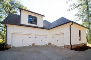 A white two-story house with a three-car garage and gabled roof, surrounded by trees. The garage doors are closed.