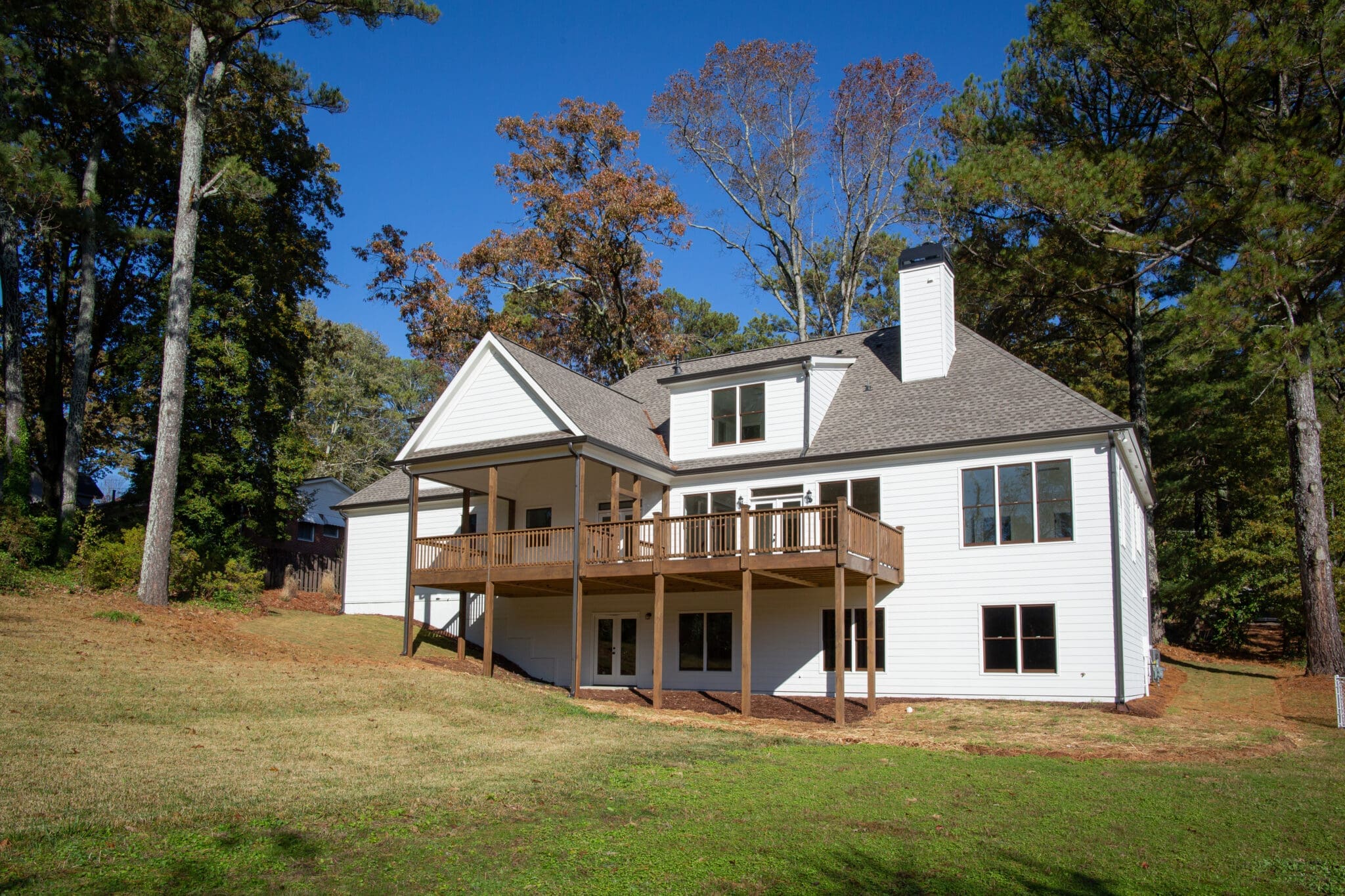 A two-story white house with a wooden deck, surrounded by trees and a grassy yard, under a clear blue sky.