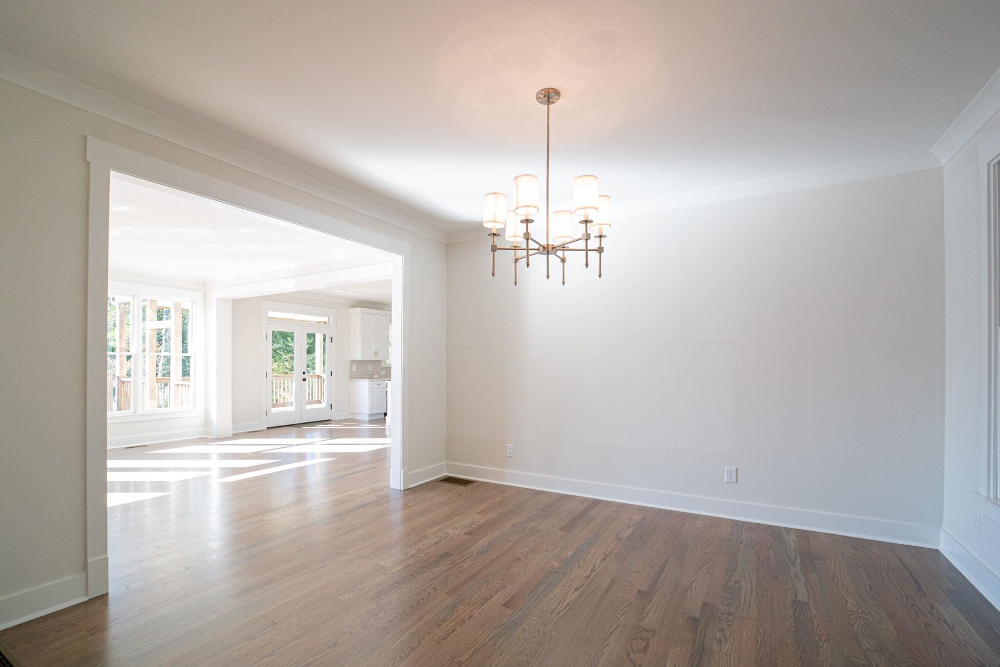A spacious, empty room with hardwood floors, white walls, and a modern chandelier. The area opens to a sunlit room with large windows and another room visible in the background.