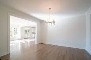 A spacious, empty room with hardwood floors, white walls, and a modern chandelier. The area opens to a sunlit room with large windows and another room visible in the background.