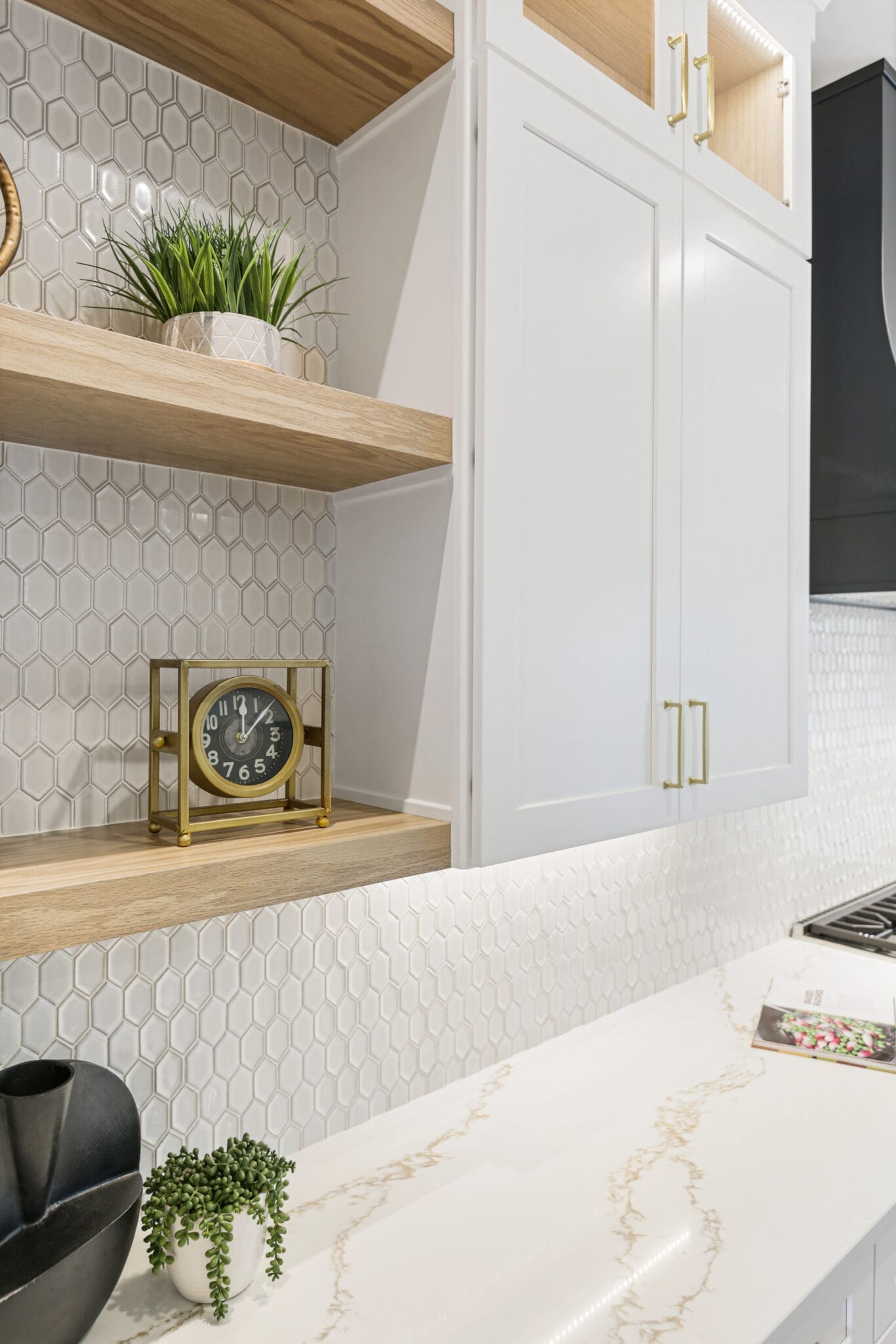 A modern kitchen with hexagonal tile backsplash, wooden and white cabinets, a brass clock on an open shelf, and a white marbled countertop. A black vase and a book are on the counter.