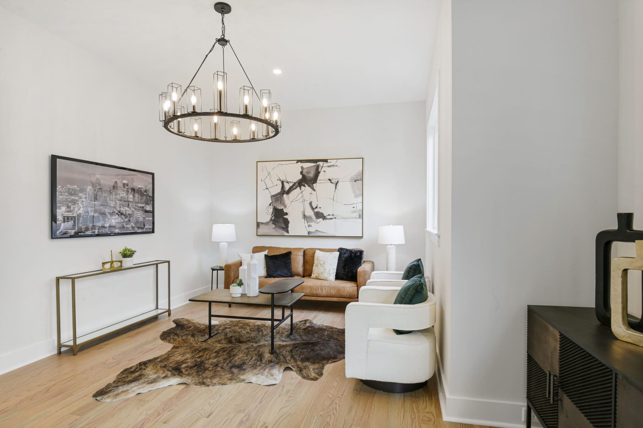 A modern living room with light-colored walls, a brown sofa, white armchair, and a cowhide rug. The room features two abstract artworks, a large window, and a chandelier with exposed bulbs.