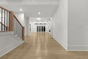 A modern, spacious hallway with light wood floors, white walls, and wooden stair railings leads to a glass door at the far end.
