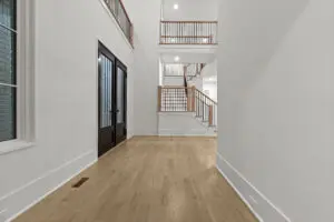 A spacious, modern foyer featuring light wood flooring, white walls, a dark double door entrance, and an open staircase with wood railing leading to an upper hallway.