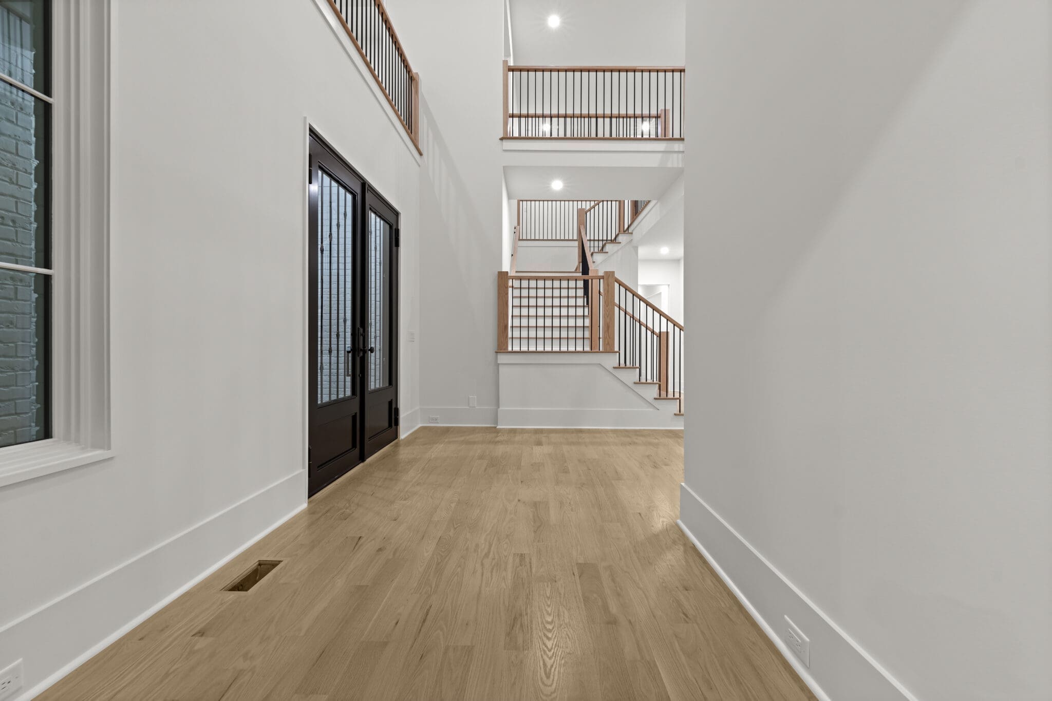 A spacious, empty hallway with light wood flooring and white walls. The hallway features a black double-door entrance and stairs with wooden railings leading to the upper level.
