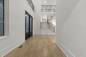 A spacious, empty hallway with light wood flooring and white walls. The hallway features a black double-door entrance and stairs with wooden railings leading to the upper level.